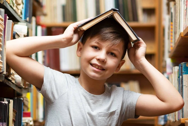Anak laki-laki tersenyum di antara rak buku sambil menutup kepala dengan buku terbuka.
