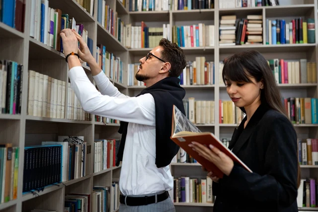 Dua mahasiswa sedang mencari dan membaca buku di rak perpustakaan kampus.