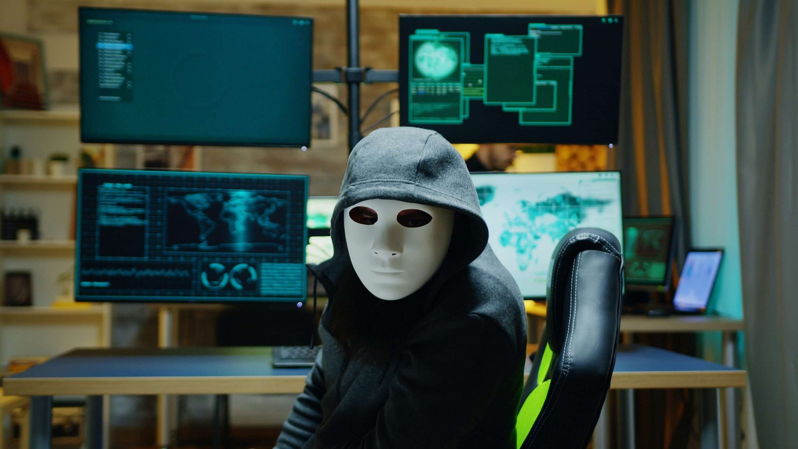 Seorang pelaku kejahatan siber bertopeng dan mengenakan hoodie duduk di ruang gelap penuh monitor sistem dunia maya